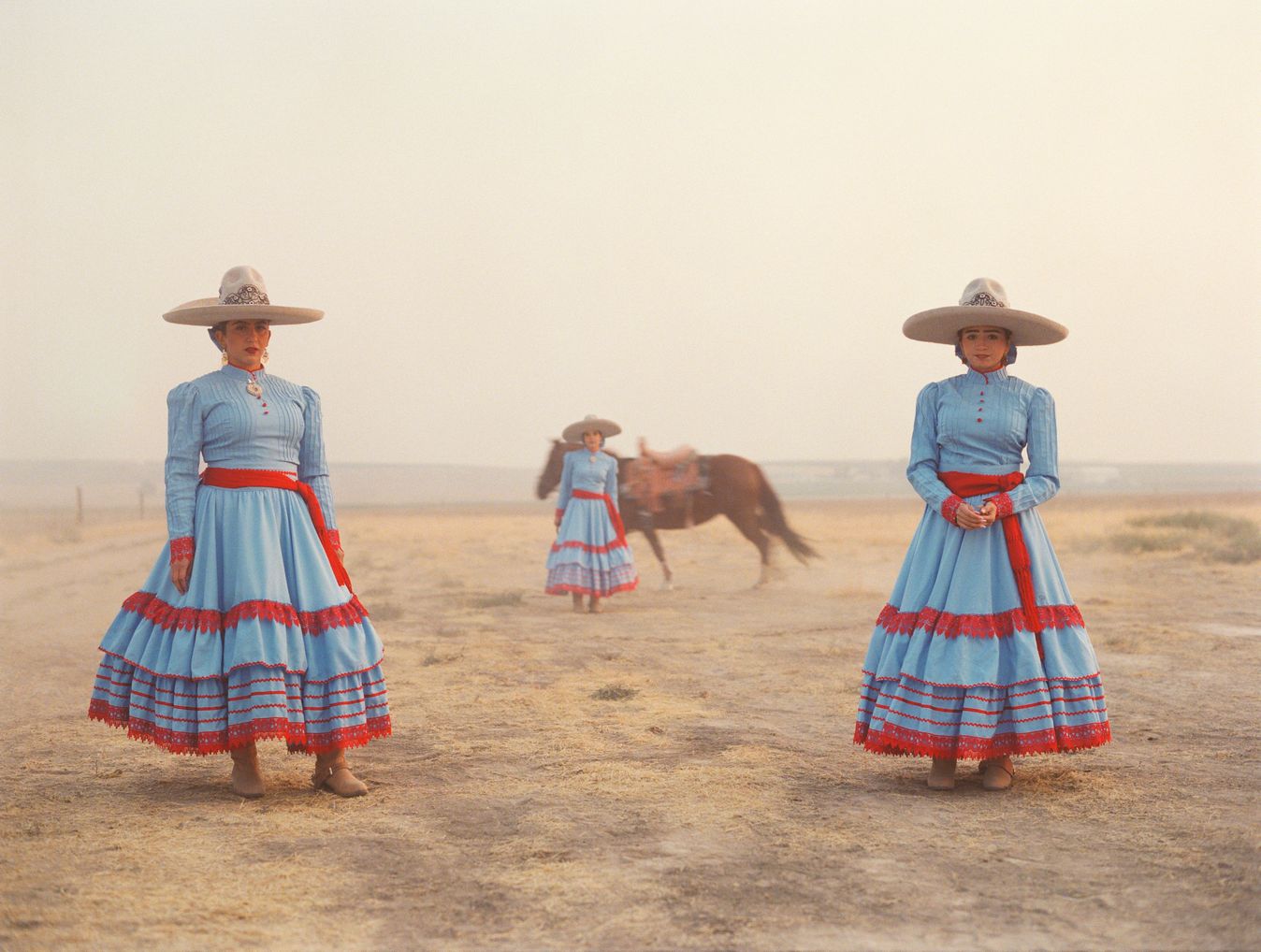 Portrait of three women in pale blue dresses with red trim and wide brimmed hats facing the camera with a horse in the background.