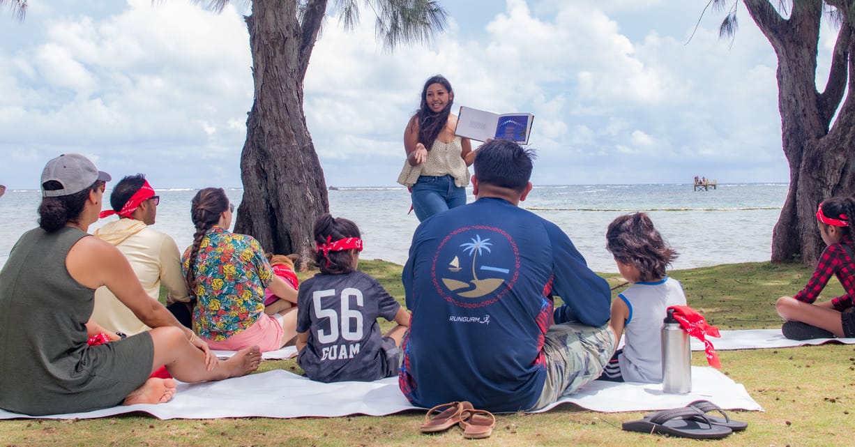 A group of people seated on a picnic blanket at a beach park, attentively listening to a standing presenter holding a book. In the background, the ocean and a clear sky are visible.