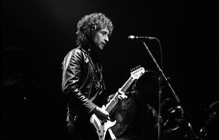 Performance image of Bob Dylan holding a guitar and standing in front of  a microphone.
