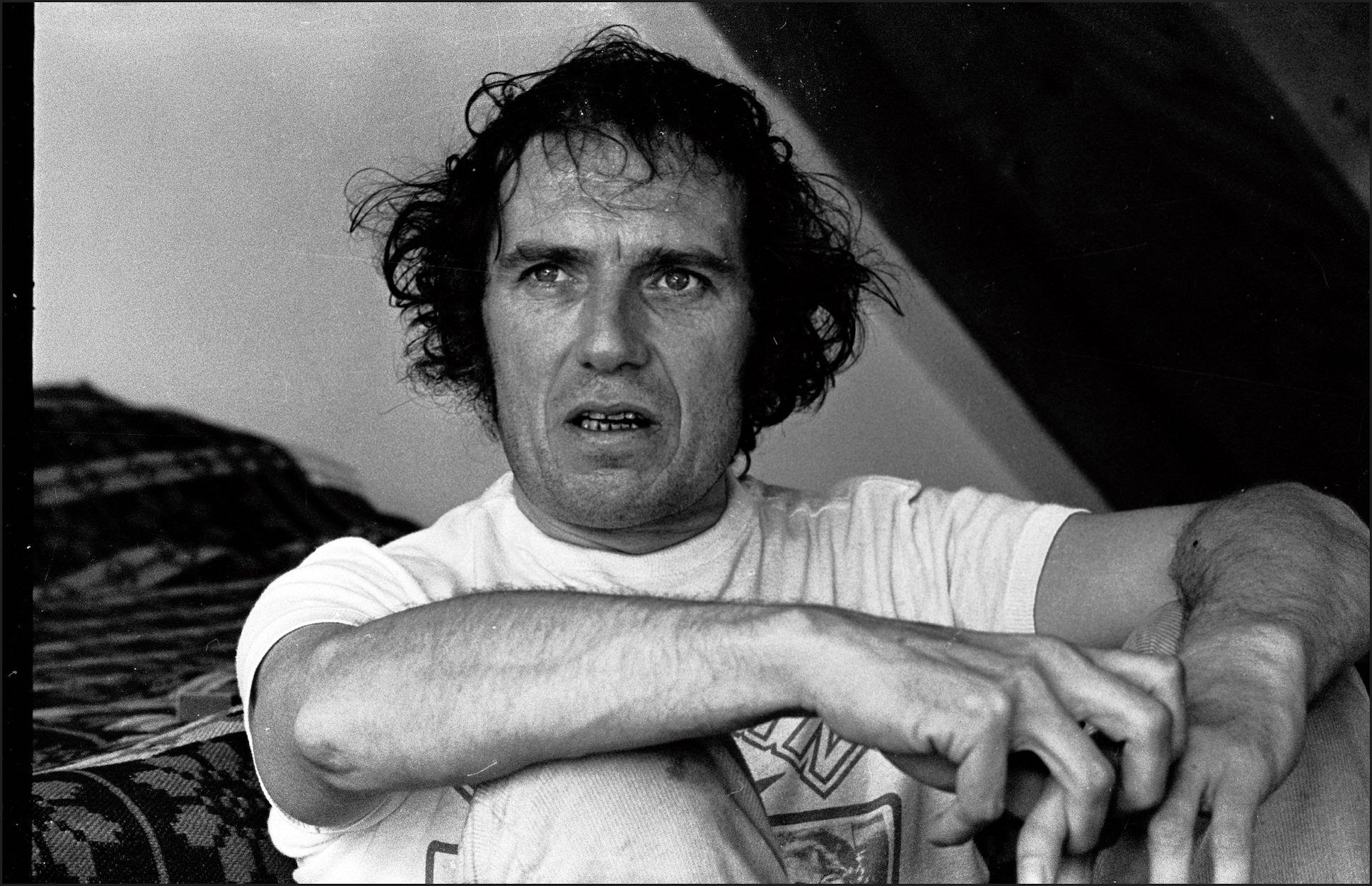Black and white portrait of poet Christopher Logue in a white t-shirt sitting on the ground.