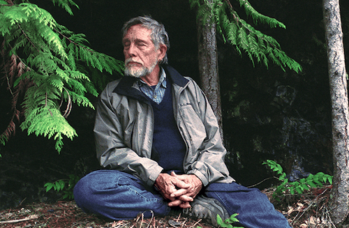 Image of Gary Snyder sitting on the ground with eyes closed in front of trees.