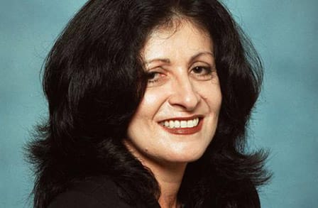 Portrait of poet Judith Ortiz Cofer with long, black hair smiling in front of a turquoise background.