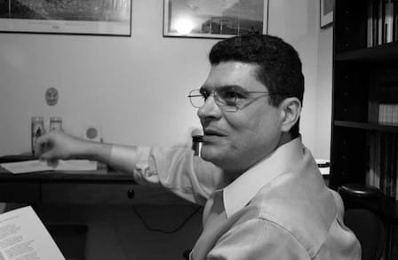 Black and white side profile of poet Orlanda Ricardo Menes in a button down shirt at his desk.