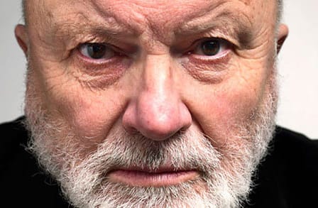 Tight-cropped portrait of Geoffrey Hill, face tilted slightly downward and staring directly into the camera.