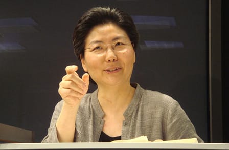 Color Photograph of Myung Mi Kim mid speech at a podium