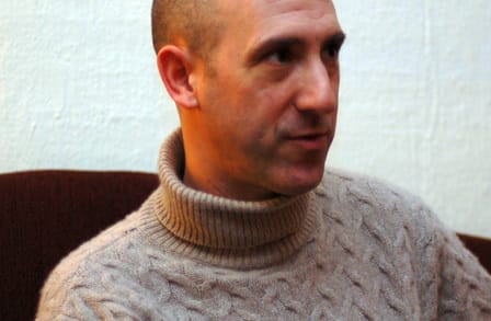 Headshot of poet Andrew Feld sitting on a maroon couch, wearing a turtleneck sweater.