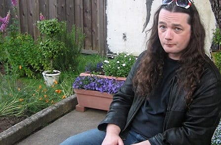 Color photograph of writer Garrett Caples sitting in a backyard surrounded by flowers