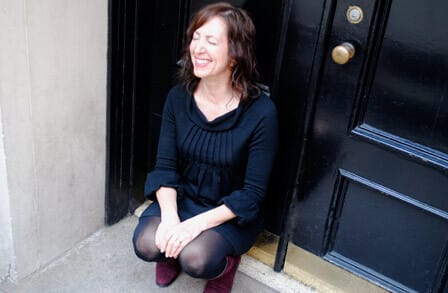 Photograph of poet Joy Katz crouching by a door with eyes closed and smiling
