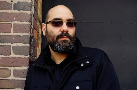 Poet Matthew Shenoda standing by a brick wall.