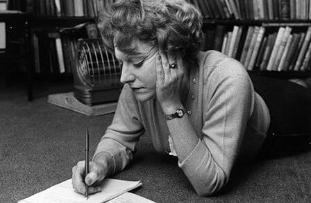 Muriel Spark lying on the carpet in front of a bookshelf, writing with a pen.