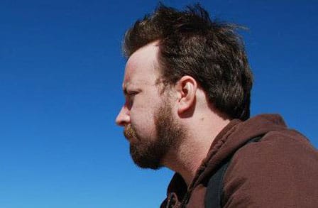Side portrait of Michael Metivier who wears a dark brown hoodie. A blue sky is in the background.