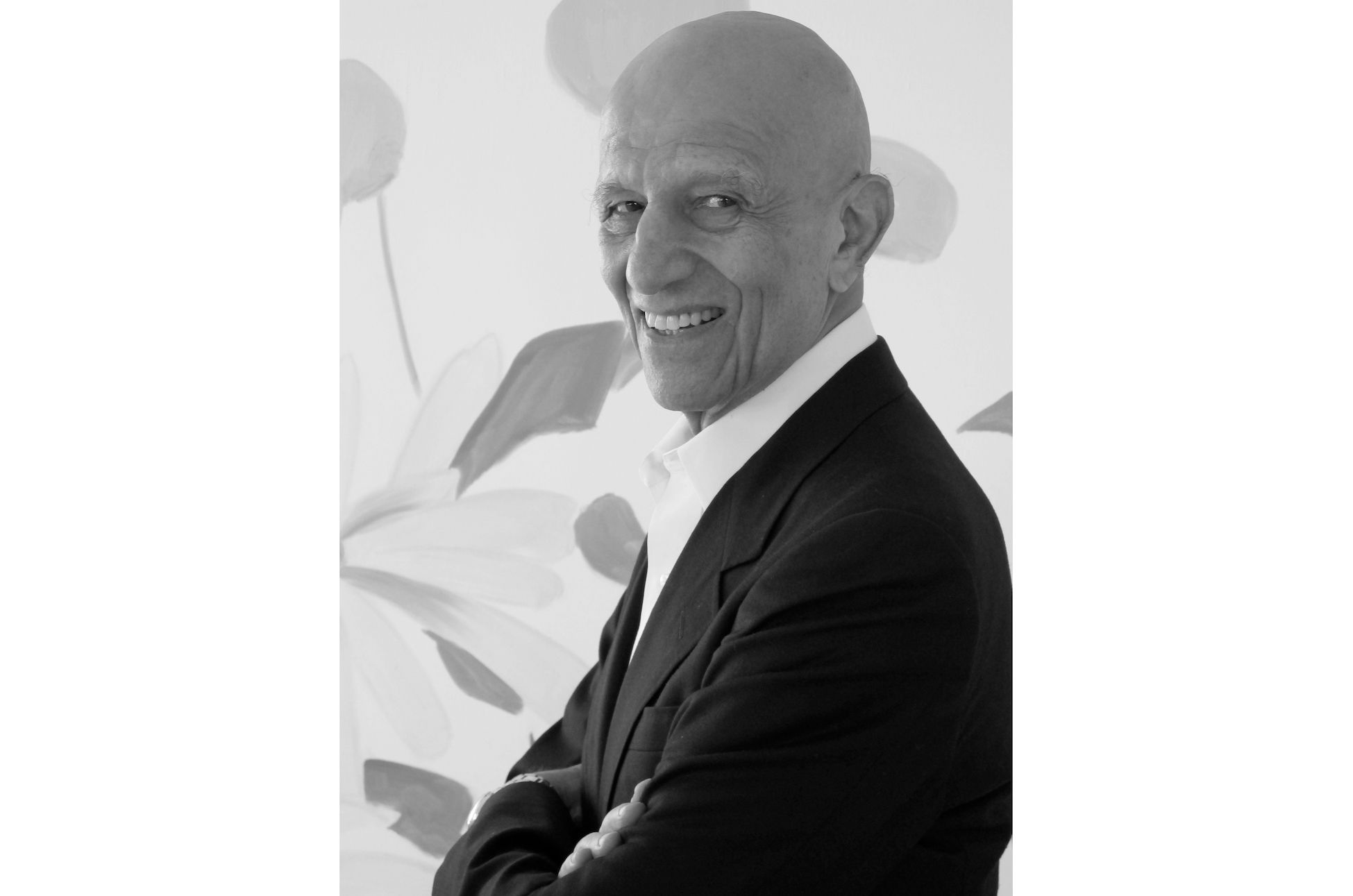 Black and white photograph of Alex Katz, smiling, wearing a dark suit and white dress shirt, and standing in front of a painting.
