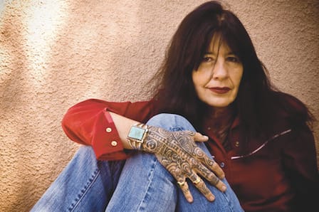 Author image of Joy Harjo sitting against a wall, wearing a red shirt.