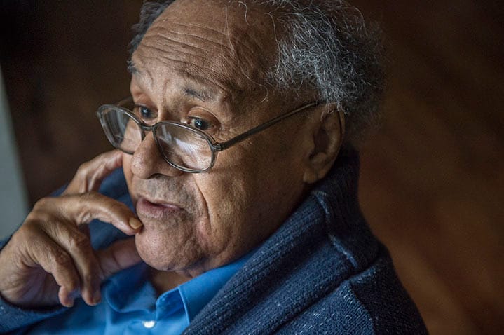 Close up angled shot of Russell Atkins's face, framed by the shawl of a navy sweater and the collar of a blue shirt. Russell is wearing glasses and gazing at something in front of him, fingers on his chin and right cheek.