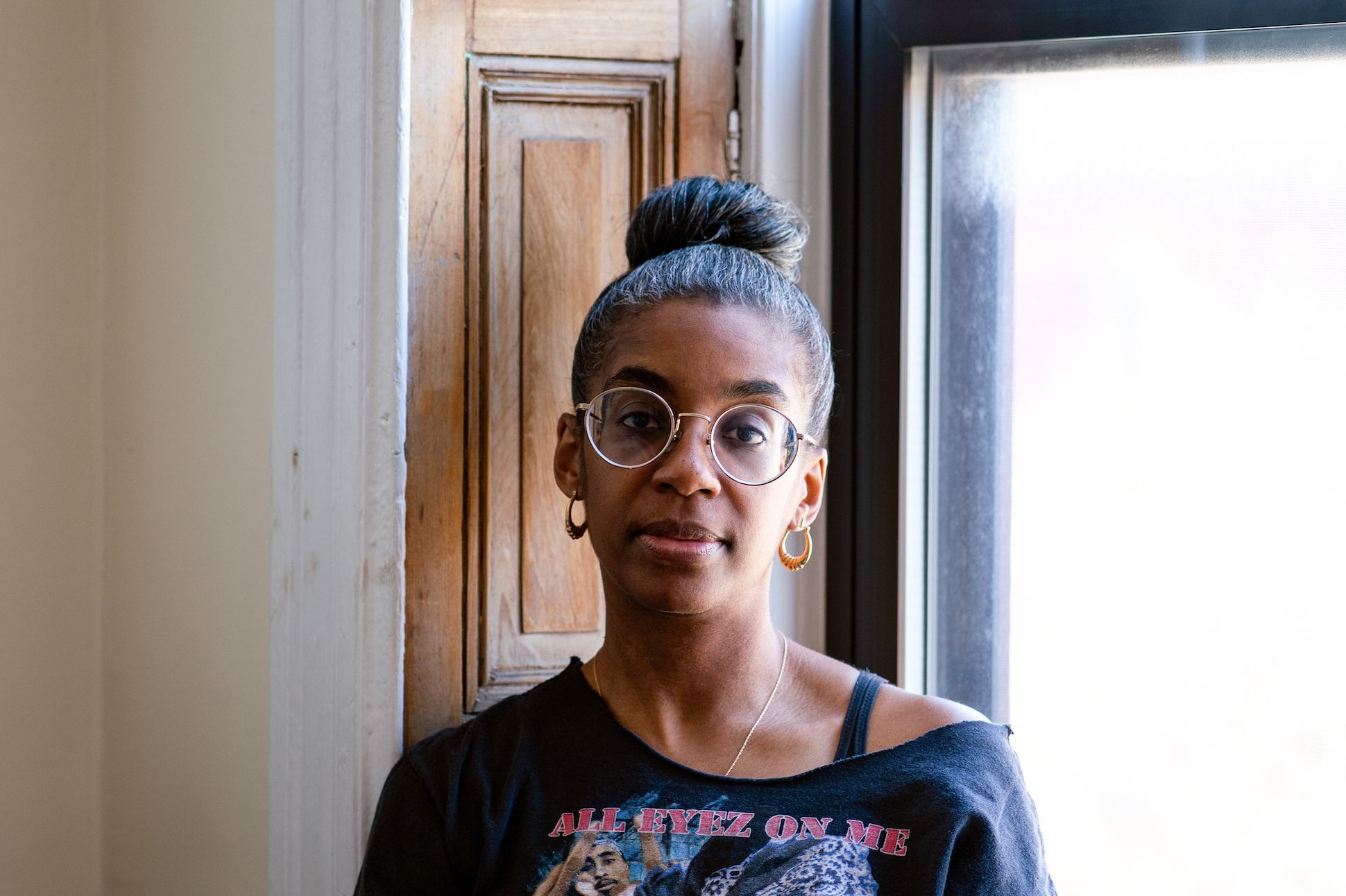 A spectacled African American woman with a hair bun and loop earrings sitting by a window