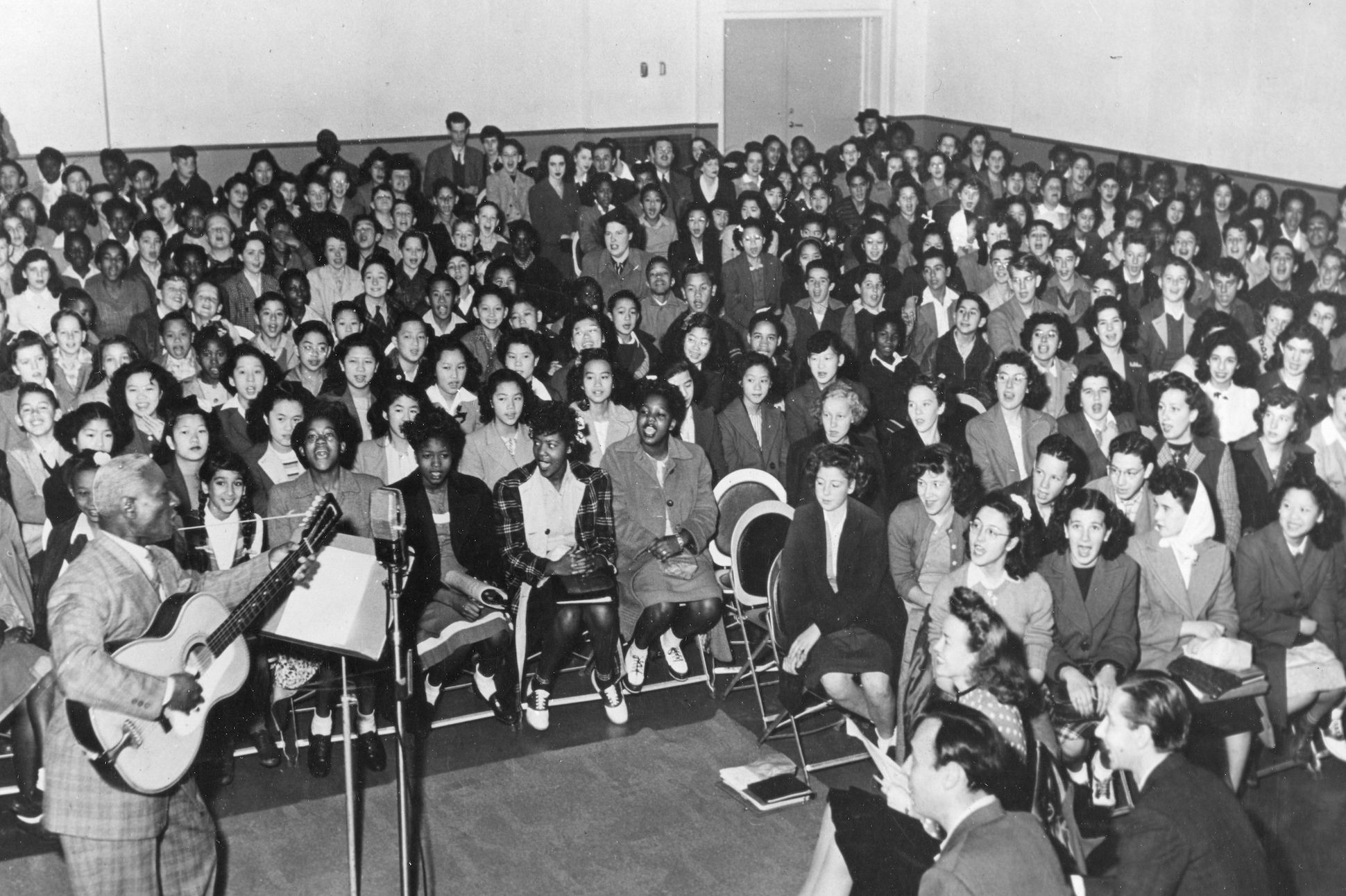 Black and white photo of Lead Belly with a guitar, surrounded by audience.