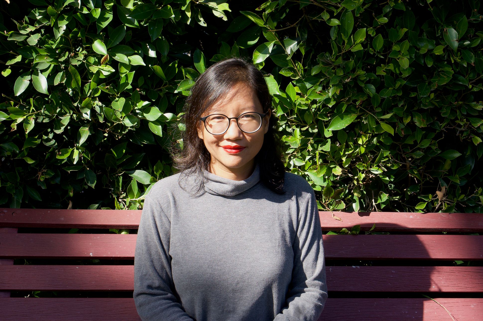 An Asian woman with glasses smiling and sitting on a red bench with brushes in the background.