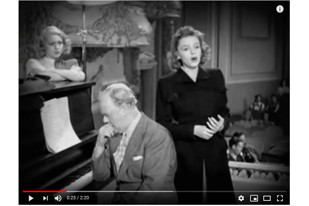 Youtube still from a black and white movie, featuring a woman and man standing next to a piano.