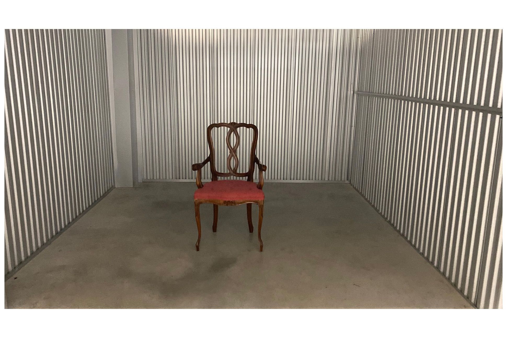 Photograph  of an upholstered wood arm chair, with red/pink cushion, in a storage unit with aluminum walls and cement floor.