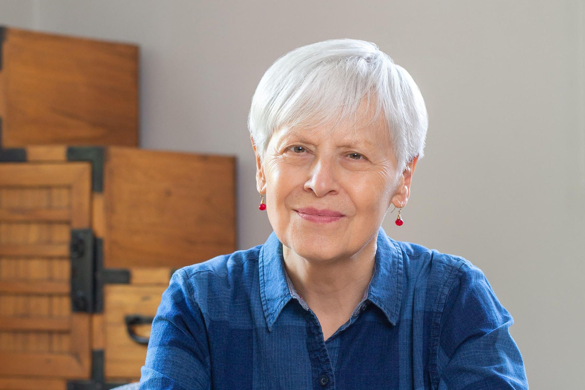 A woman with short grey hair, red earrings, and blue shirt smiling with boxes in the background.