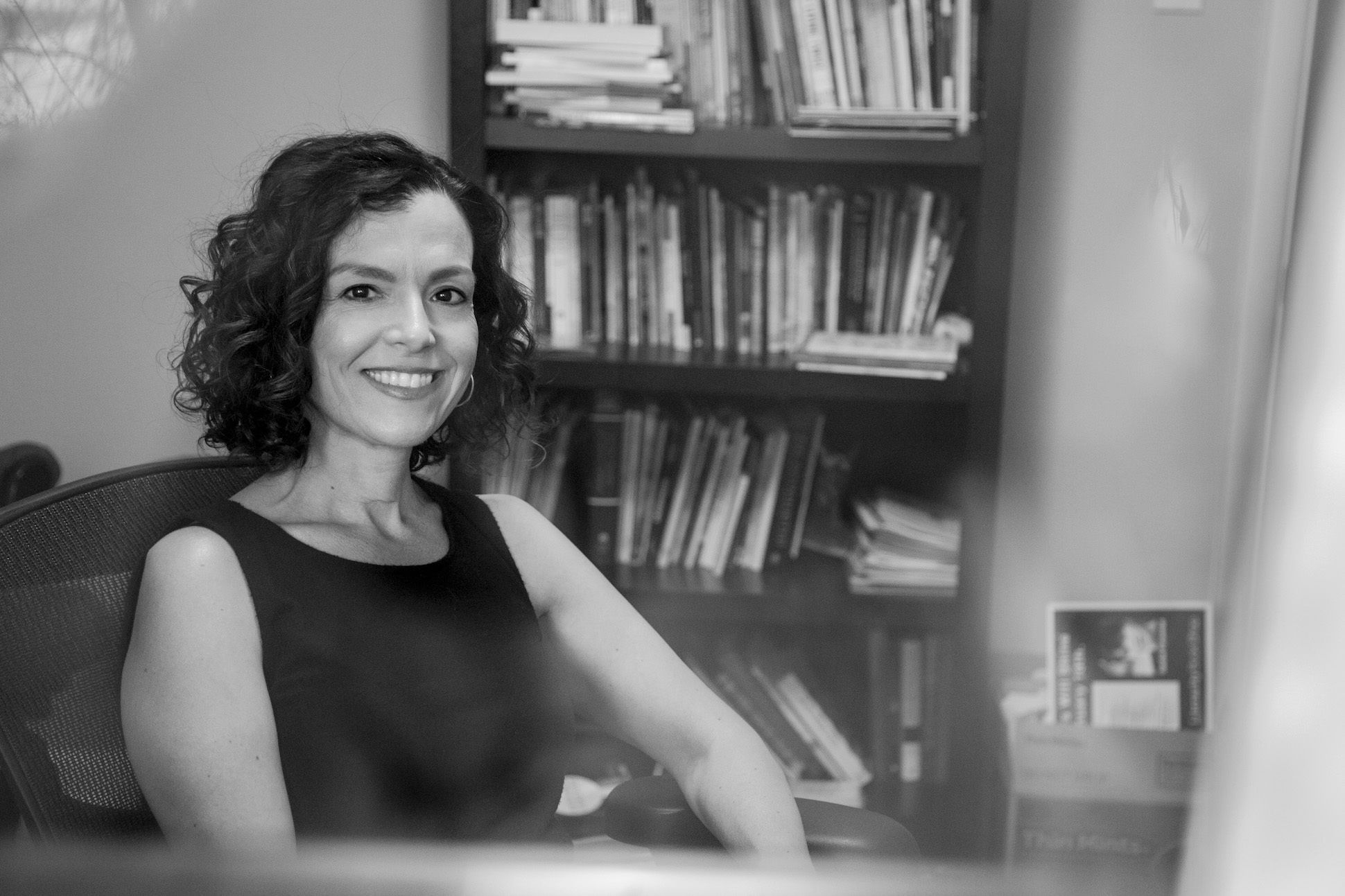 Black and white portrait of a woman with wavy hair in a black tank top sitting on a chair next to a bookshelf smiling