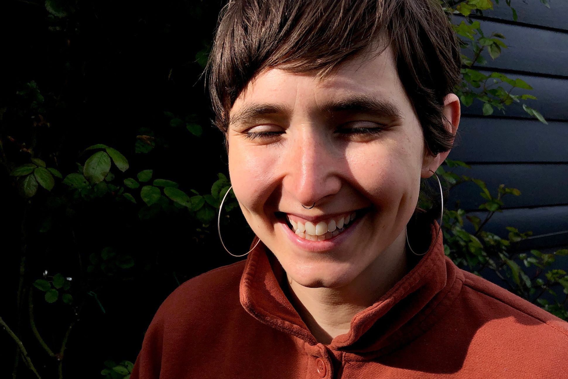 A white woman with pixie hair, hoop earrings, and an orange top is smiling in front of the exterior of a house.