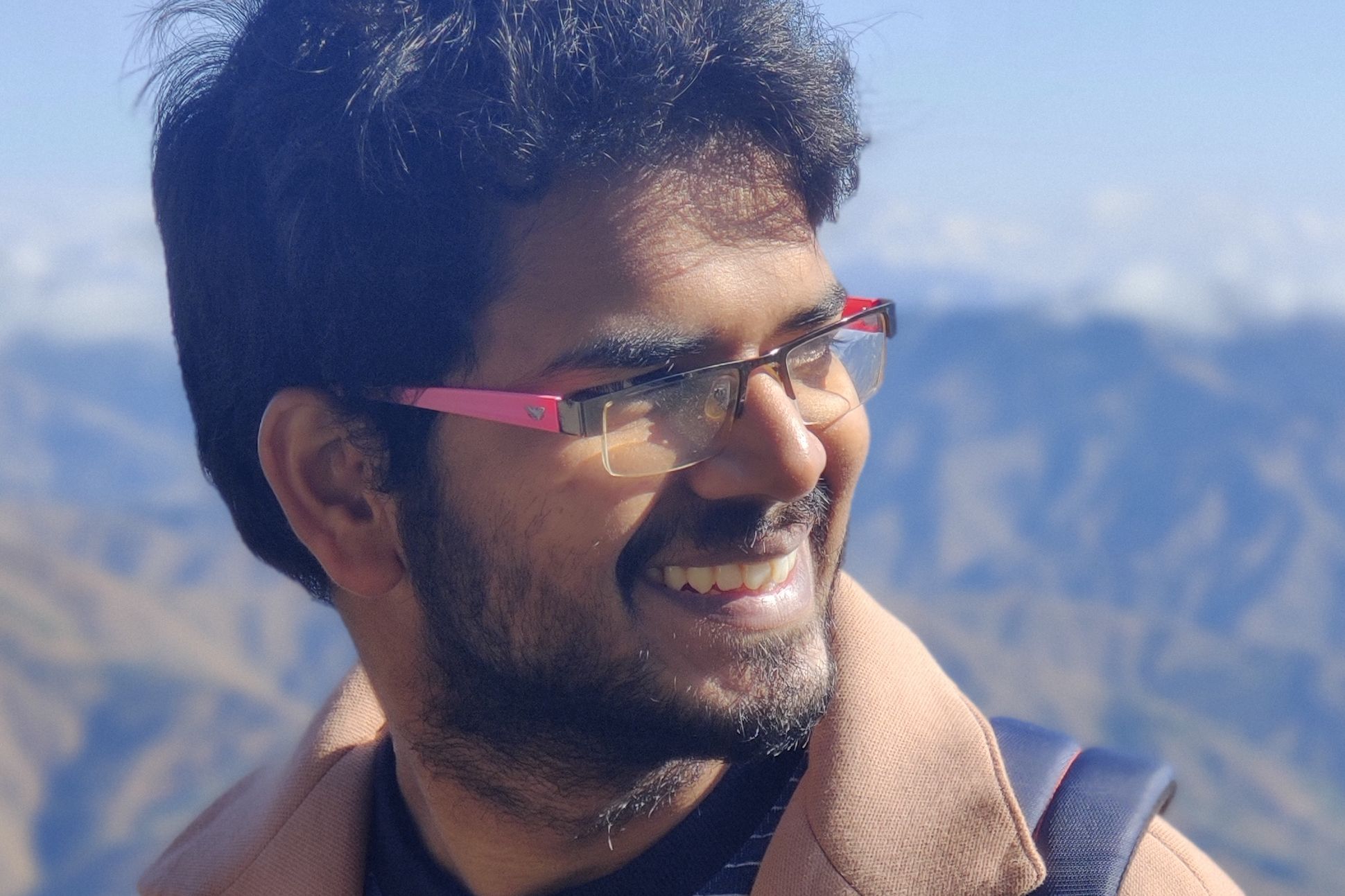An Indian male with pink glasses and beard wearing a jacket and carrying a backpack. The background is a range of mountains.