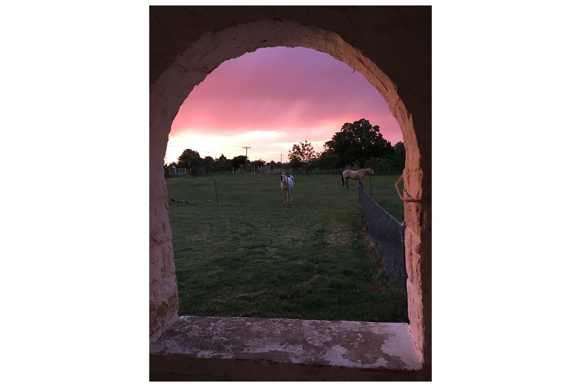 View through a stone archway of a field with horses, sunset in the background.