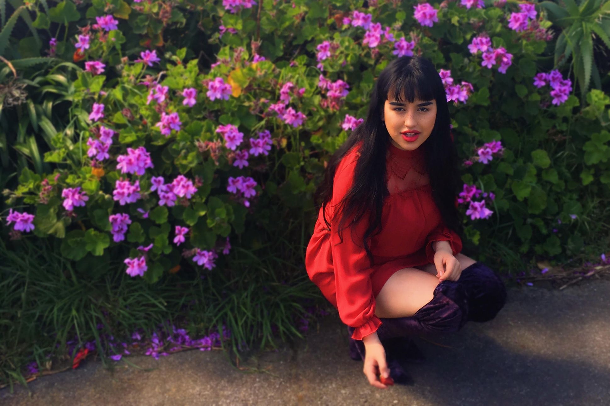 Image of Tayi Tibble wearing a red dress squatting down to pose in front of purple flowers