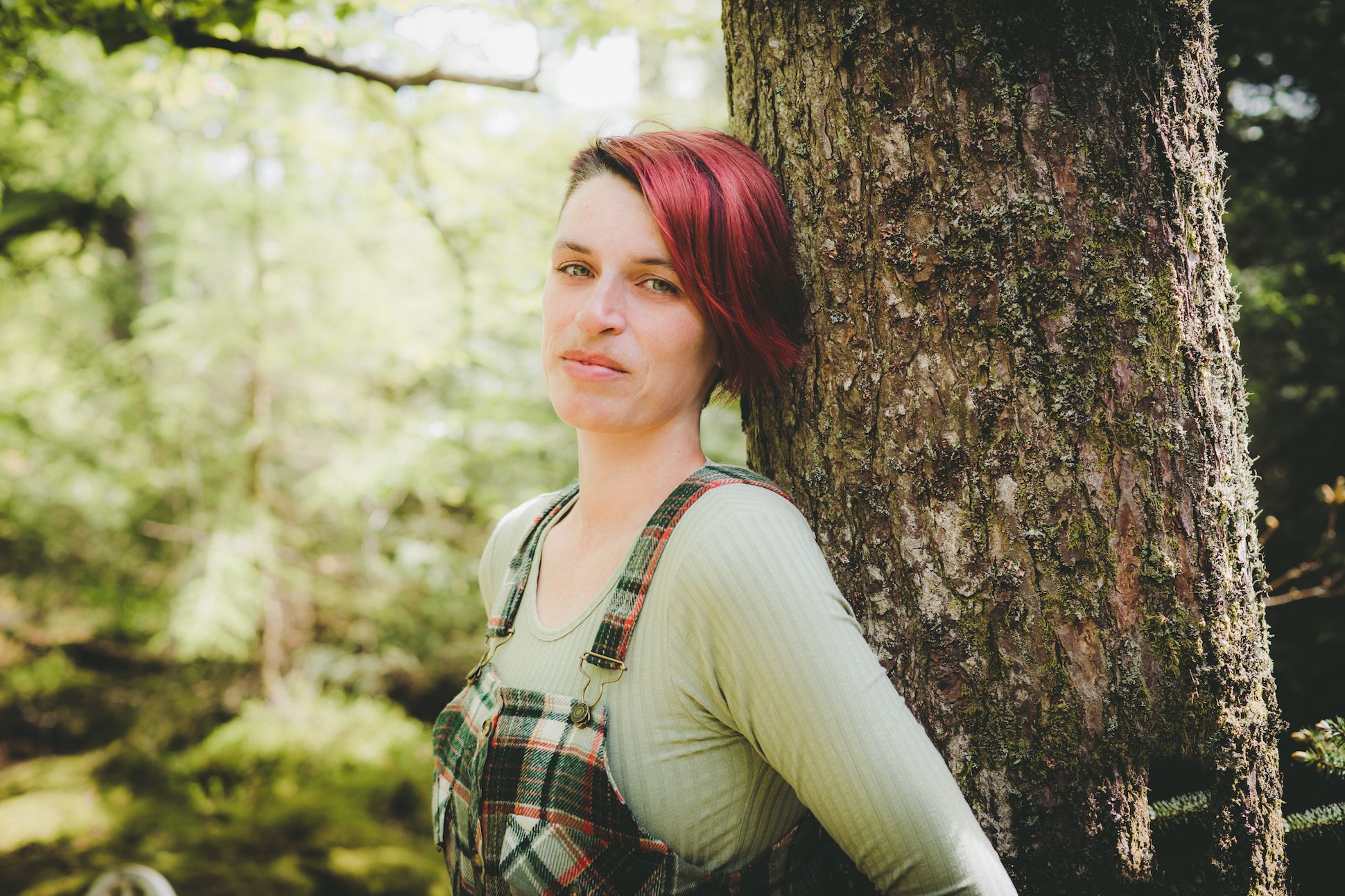 A white woman with short red hair wearing a green pullover and a plaid overalls standing by a tree. The background is bright green.