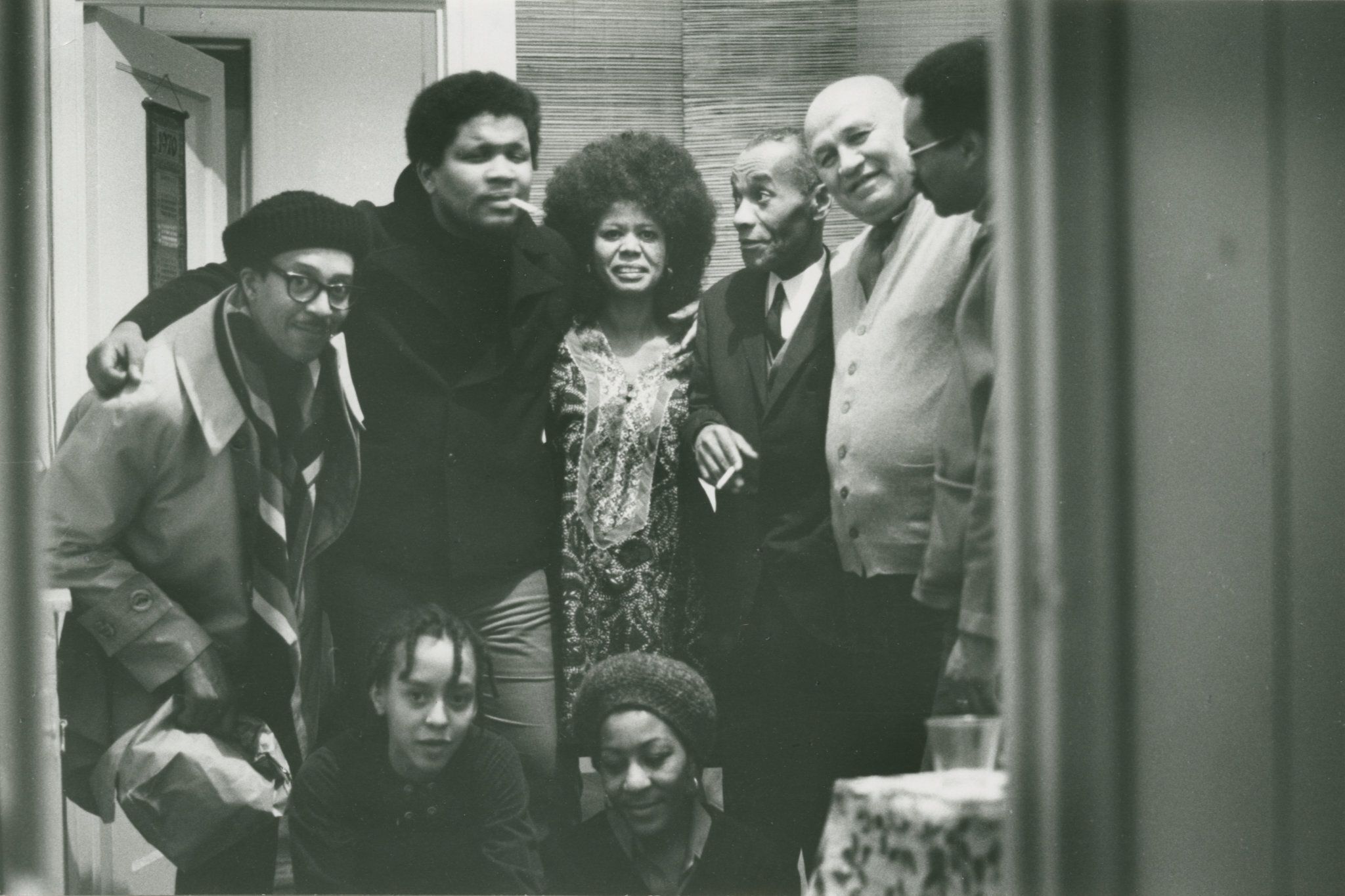 Jayne Cortez stands in the middle of other Black artists and writers, posing for the photo. Some look at the camera and others at each other. There is a table with cups slightly visible in the foreground and cracked door in the background.