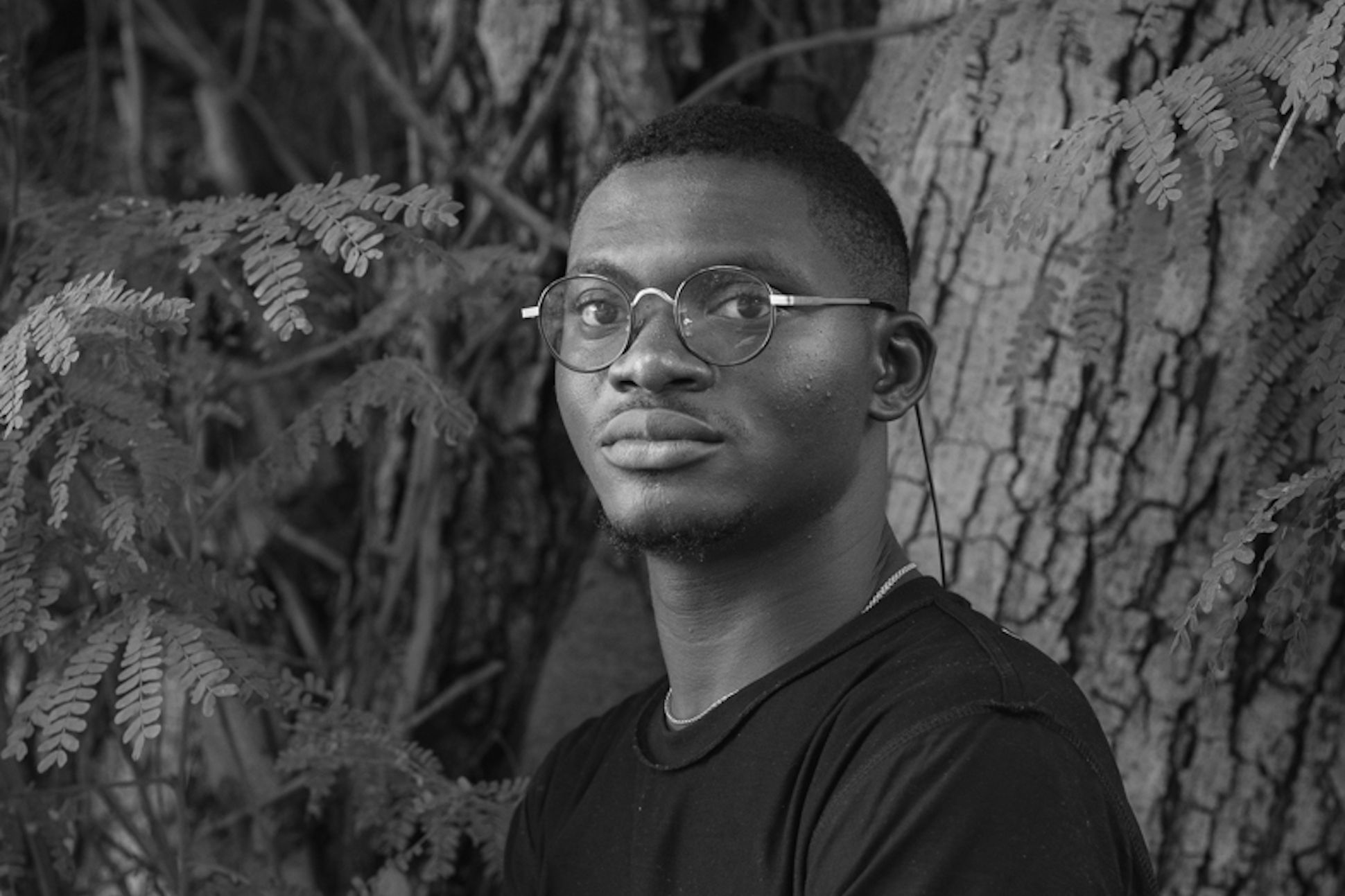 A black male with glasses and black t-shirt standing in front of trees