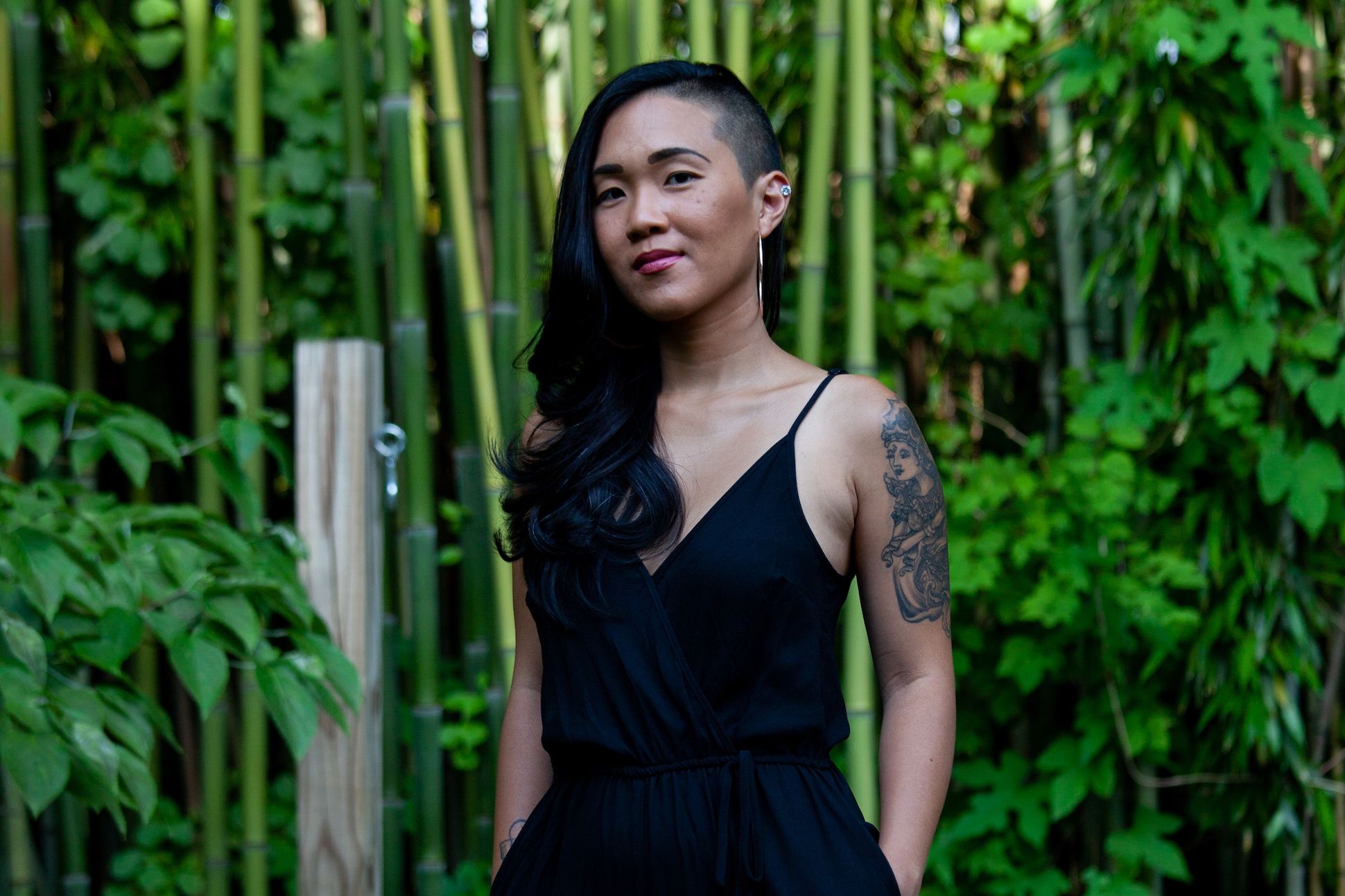 An Asian woman with one side of her long hair shaved is standing in front of bamboo trees