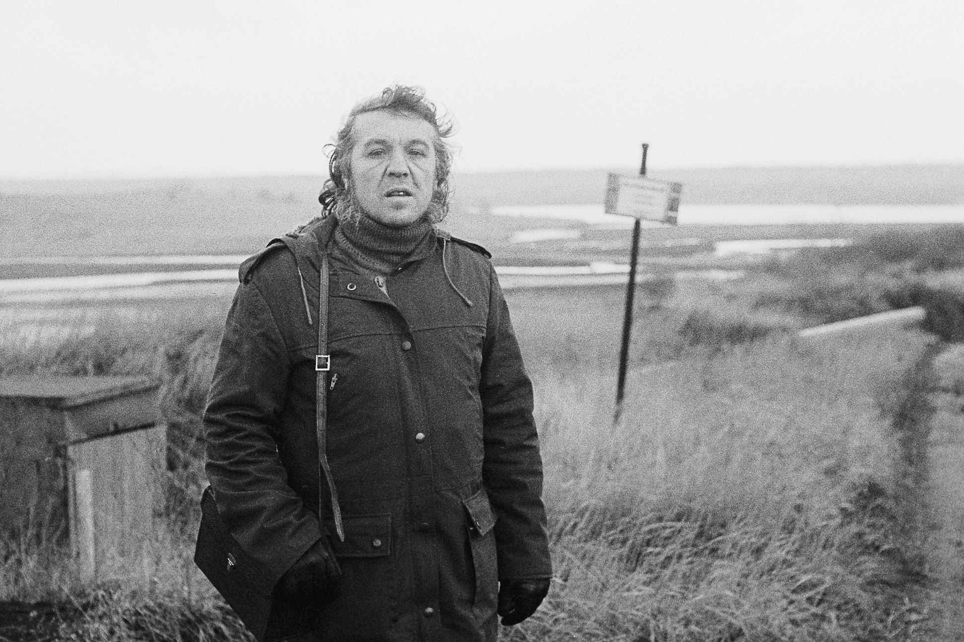 A black-and-white photograph of Wolfgang Hilbig in a dark coat and gloves, a satchel over his shoulder. Behind him is a desolate country landscape.