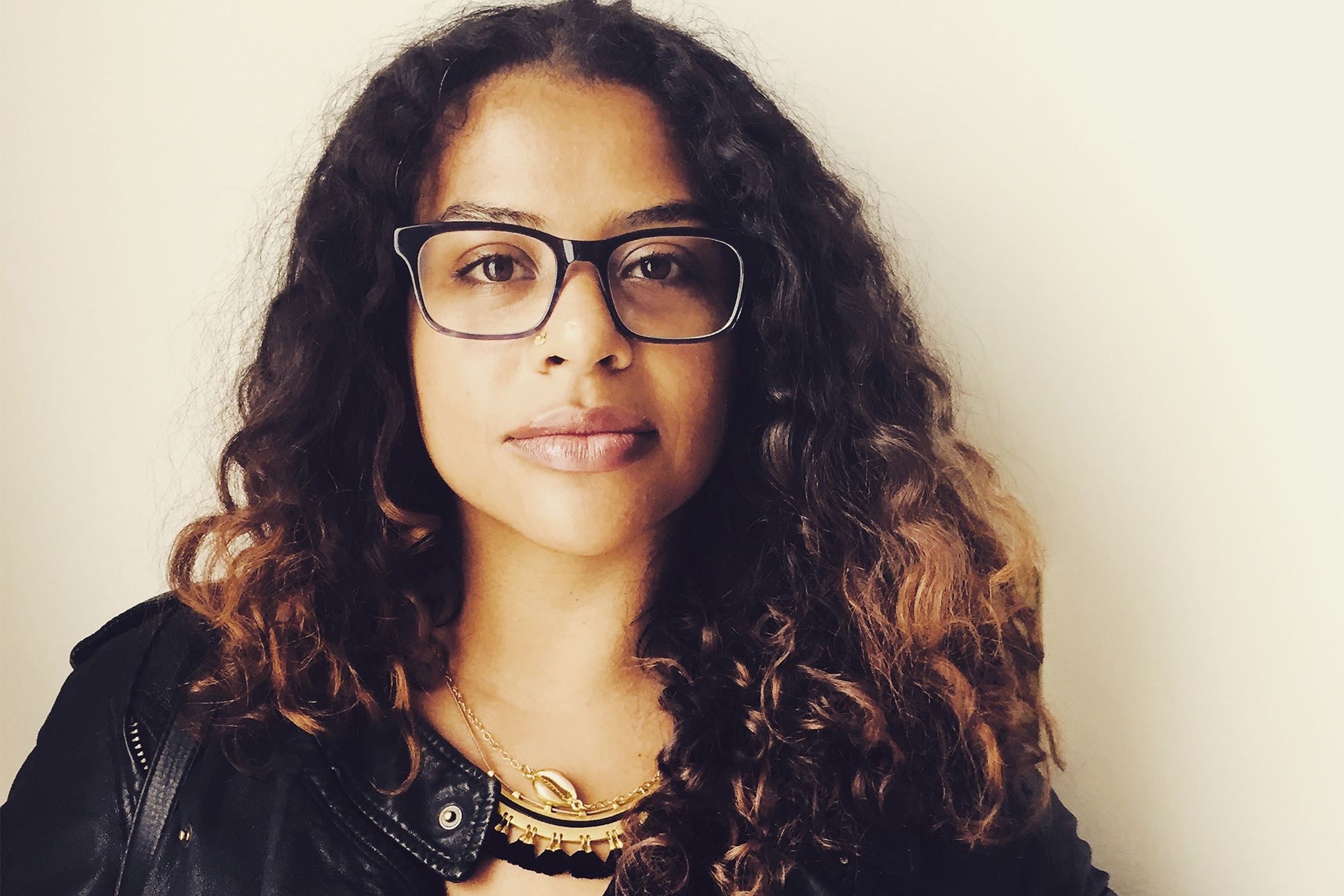 Portrait of Rio Cortez looking into the camera. She wears glasses, a gold necklace, and a leather jacket.