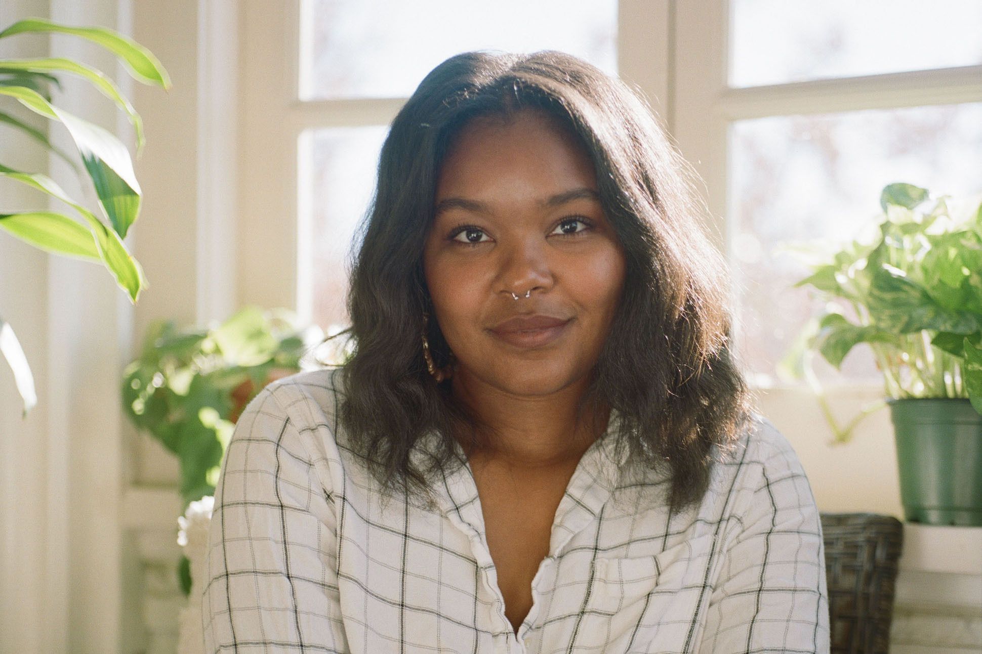 A black woman with shoulder length hair, a nose ring, and a white plaid shirt sitting in front of a window with plants.