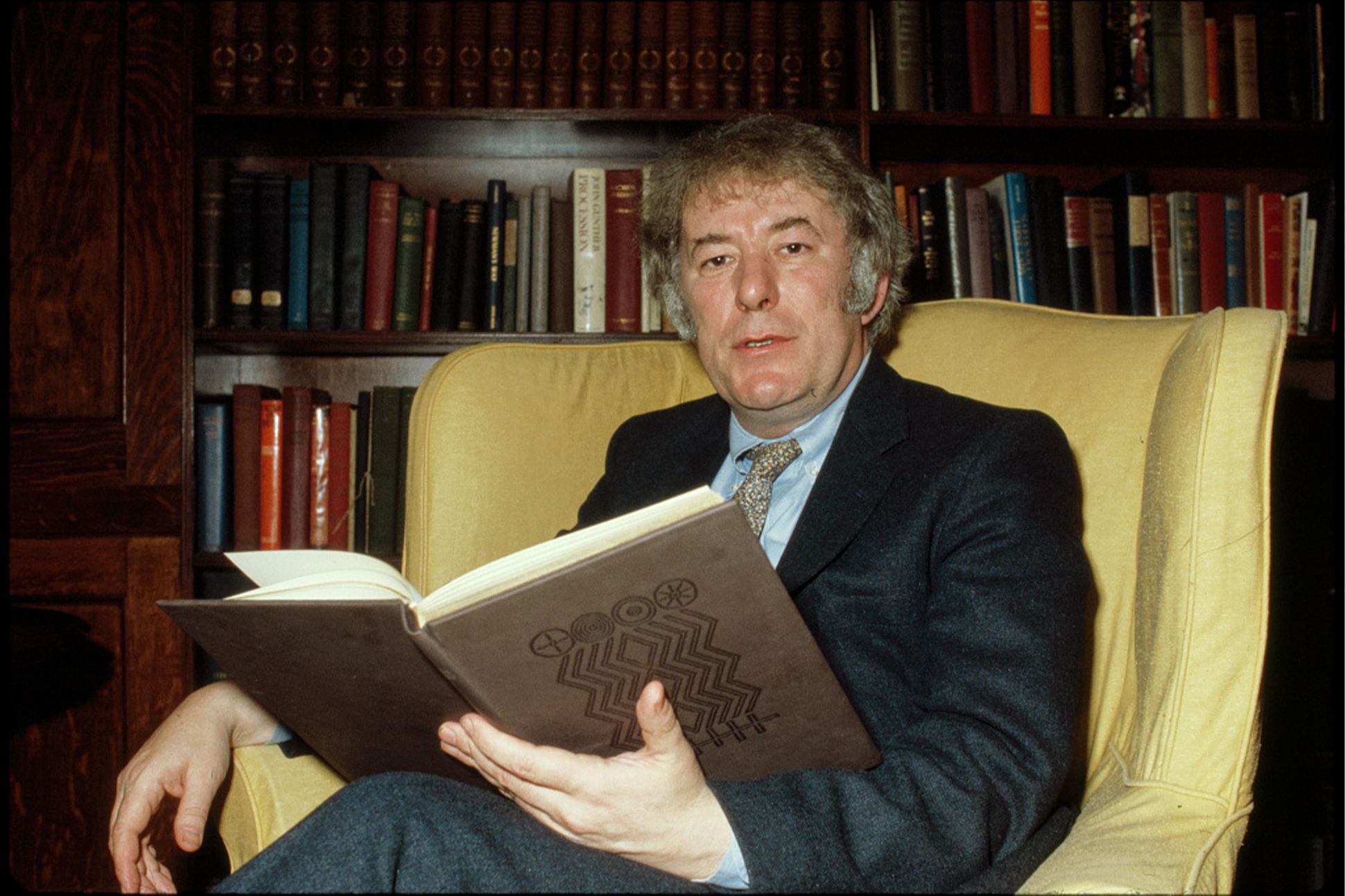 A photograph of Seamus Heaney sitting in a yellow chair, holding an open book, with bookshelves behind him.