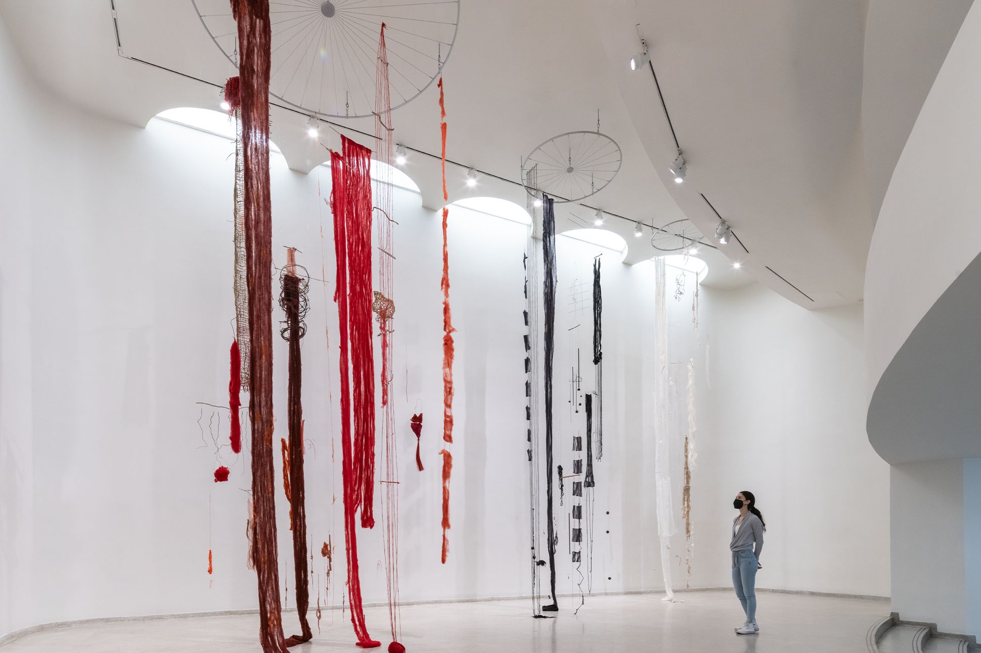 A masked museum patron stands in front of large, knotted, fiber sculptures that hang from the ceiling.