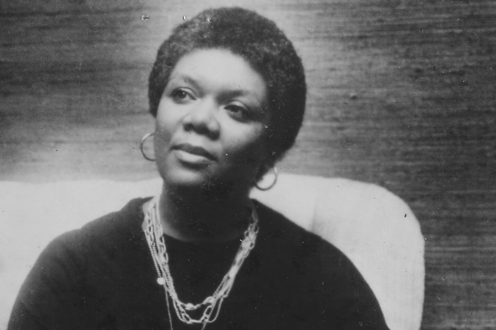 A black and white photograph of Lucille Clifton, a Black woman with a short afro, looking to the side. She is wearing a black top, hoop earrings, and a chain necklace of several strands. There is a sofa back and a wall with textured wallpaper behind her.