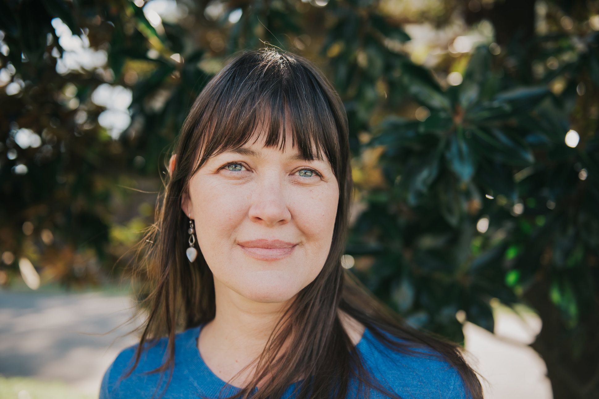 Long haired White woman in a blue top smiling in front of a tree