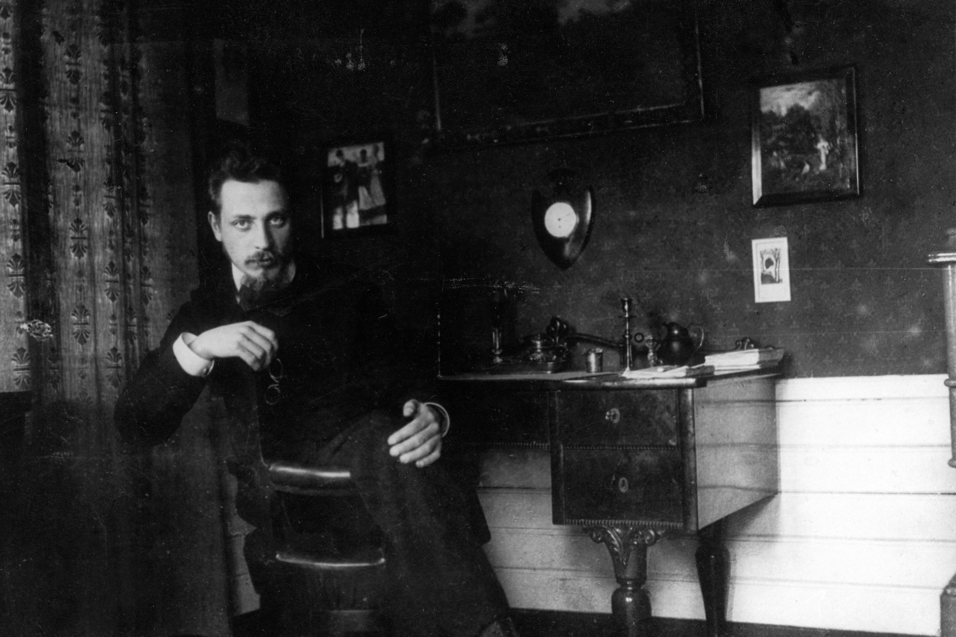 A black-and-white photograph of Rainer Maria Rilke sitting at a desk in his study, facing the camera.