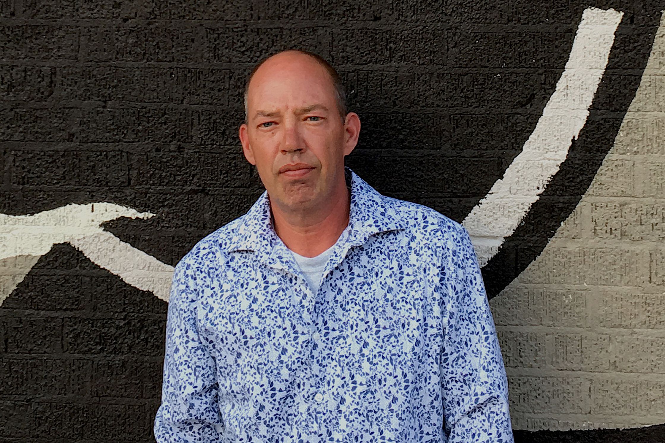 Image of poet Steve Bellin Oka posing in front of a gray mural.
