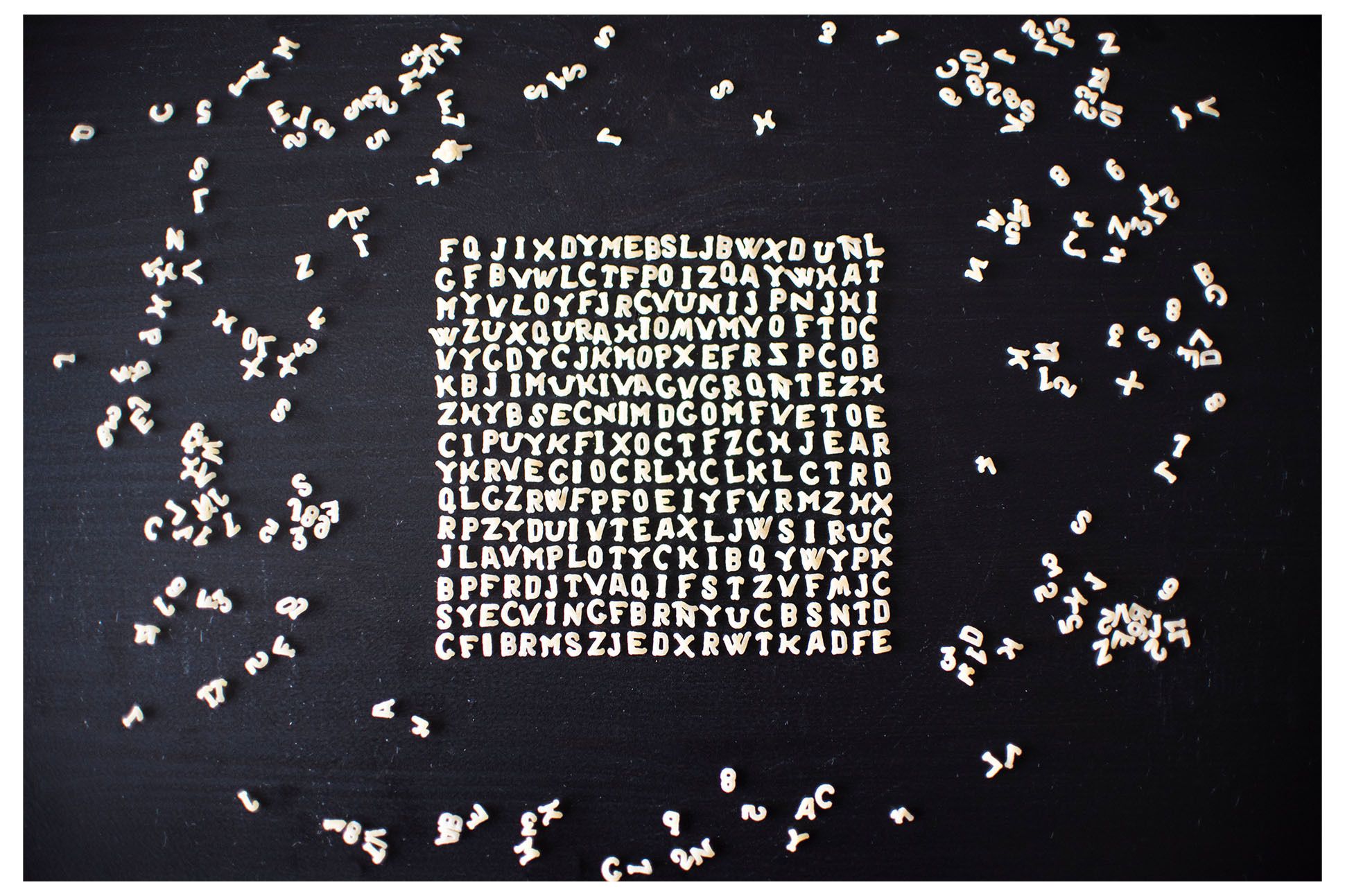 white three dimensional alphabet pieces arranged, in no discernible order, in a square shape, with random alphabet around it, against a black backdrop.