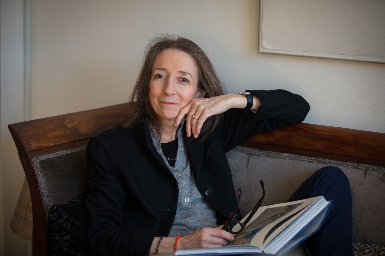 Saskia Hamilton sits with a book open in her lap, looking softly at the camera with her face resting against her left hand and her glasses in her right hand.
