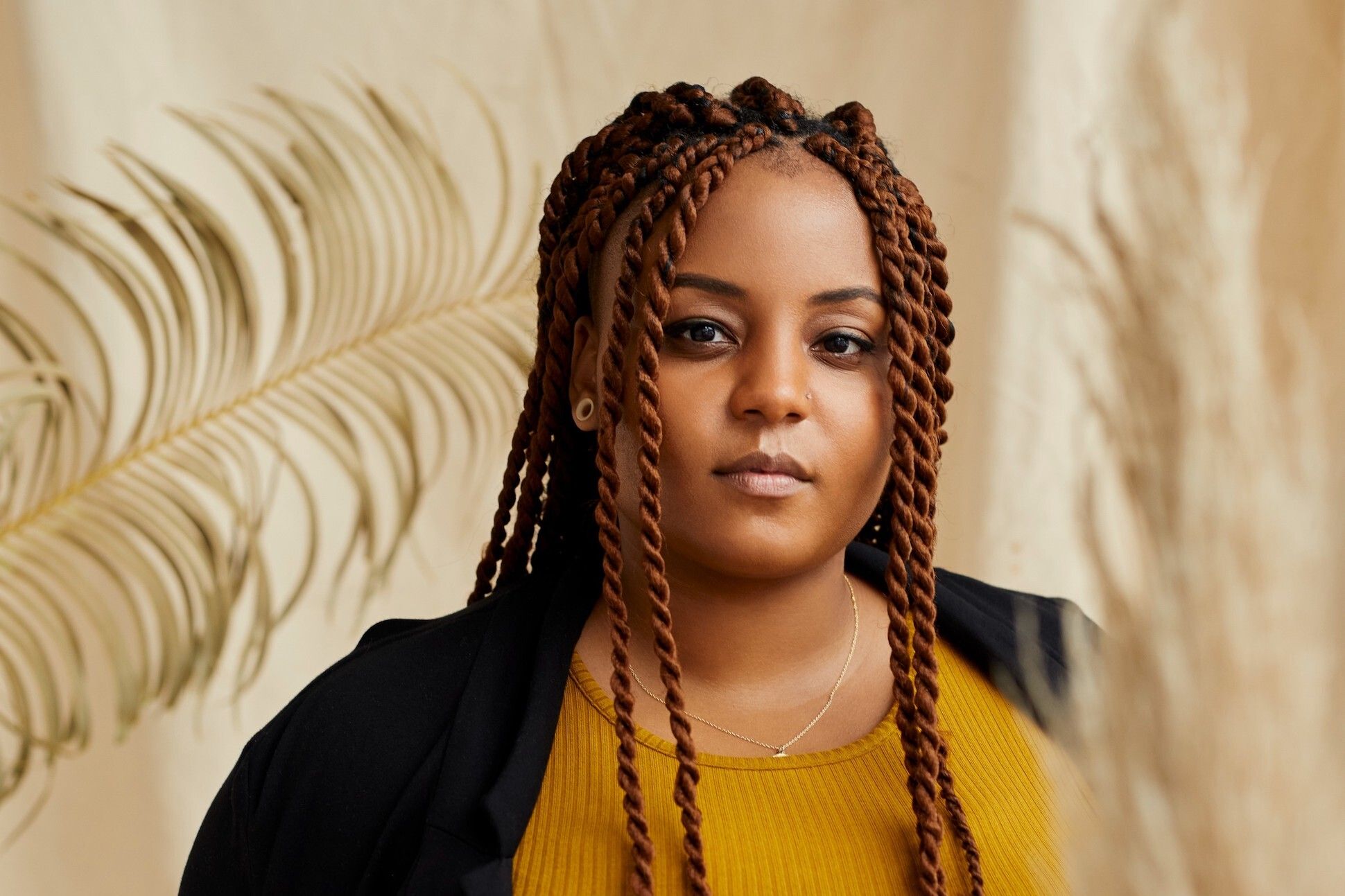 A black woman with long braided hair wearing a yellow top and a black cardigan. There is a plant with big leaf in the cream yellow background.