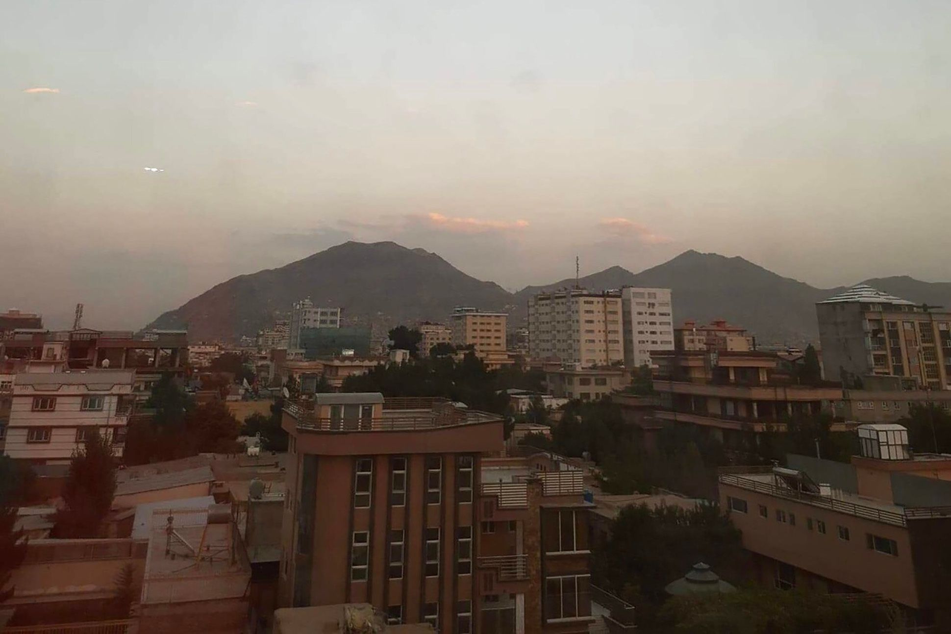 Brown, white, and beige buildings in the foreground with tall mountains in the background and low, white clouds behind the mountains. The sky is very pale blue with some soft pink accents.