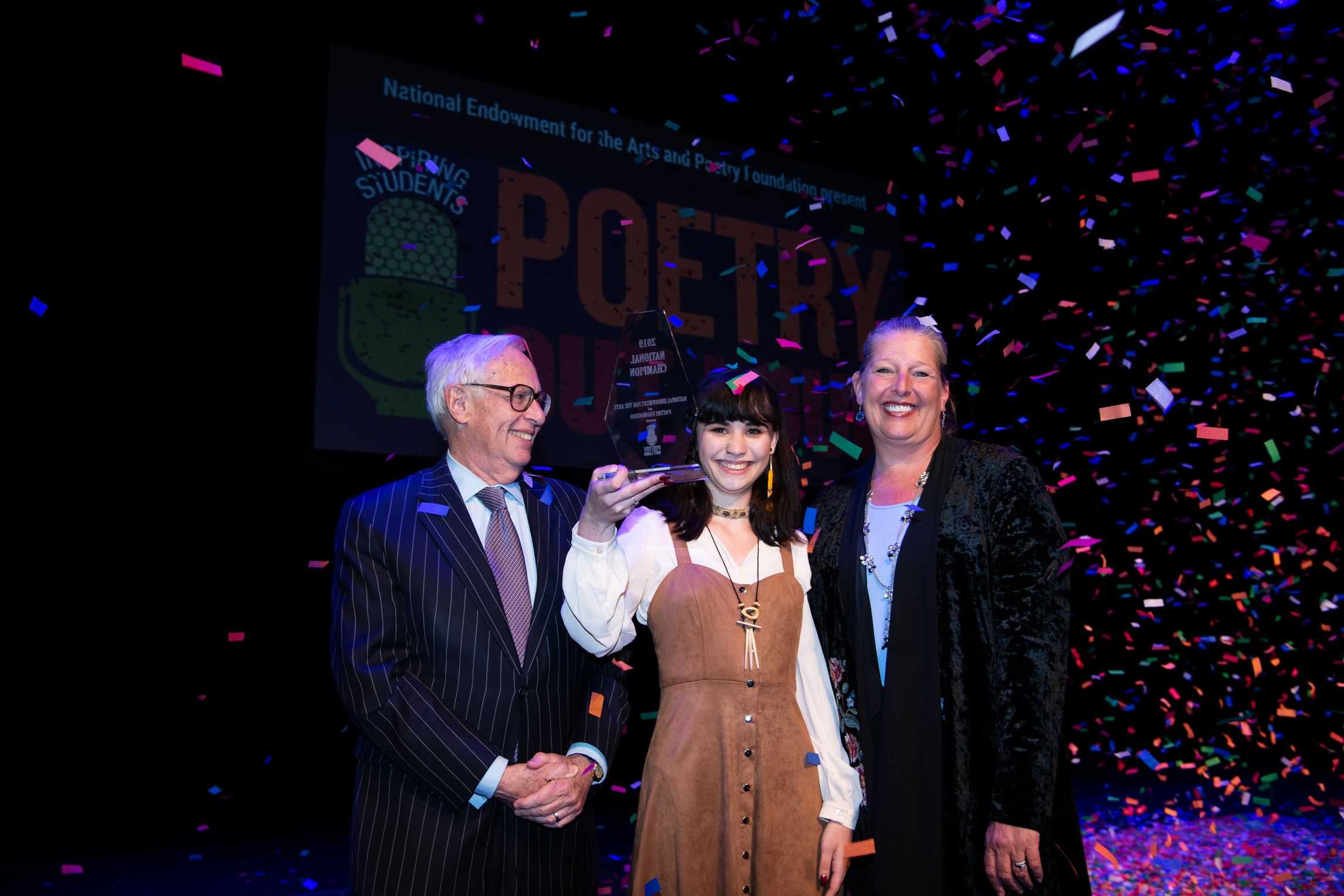 Three people (Henry Bienen, man with white hair and glasses; Isabella Callery, woman with dark hair, holding award; Mary Anne Carter, woman with blonde hair) all smiling as confetti falls around them.