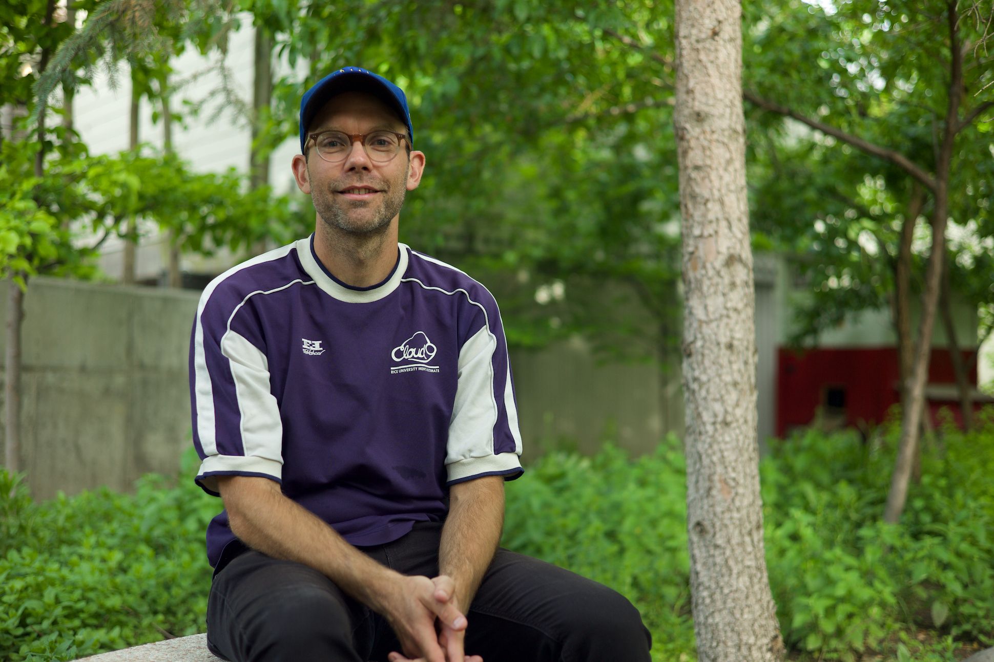 A white male wearing a purple t-shirt and a baseball cap sitting in the park.