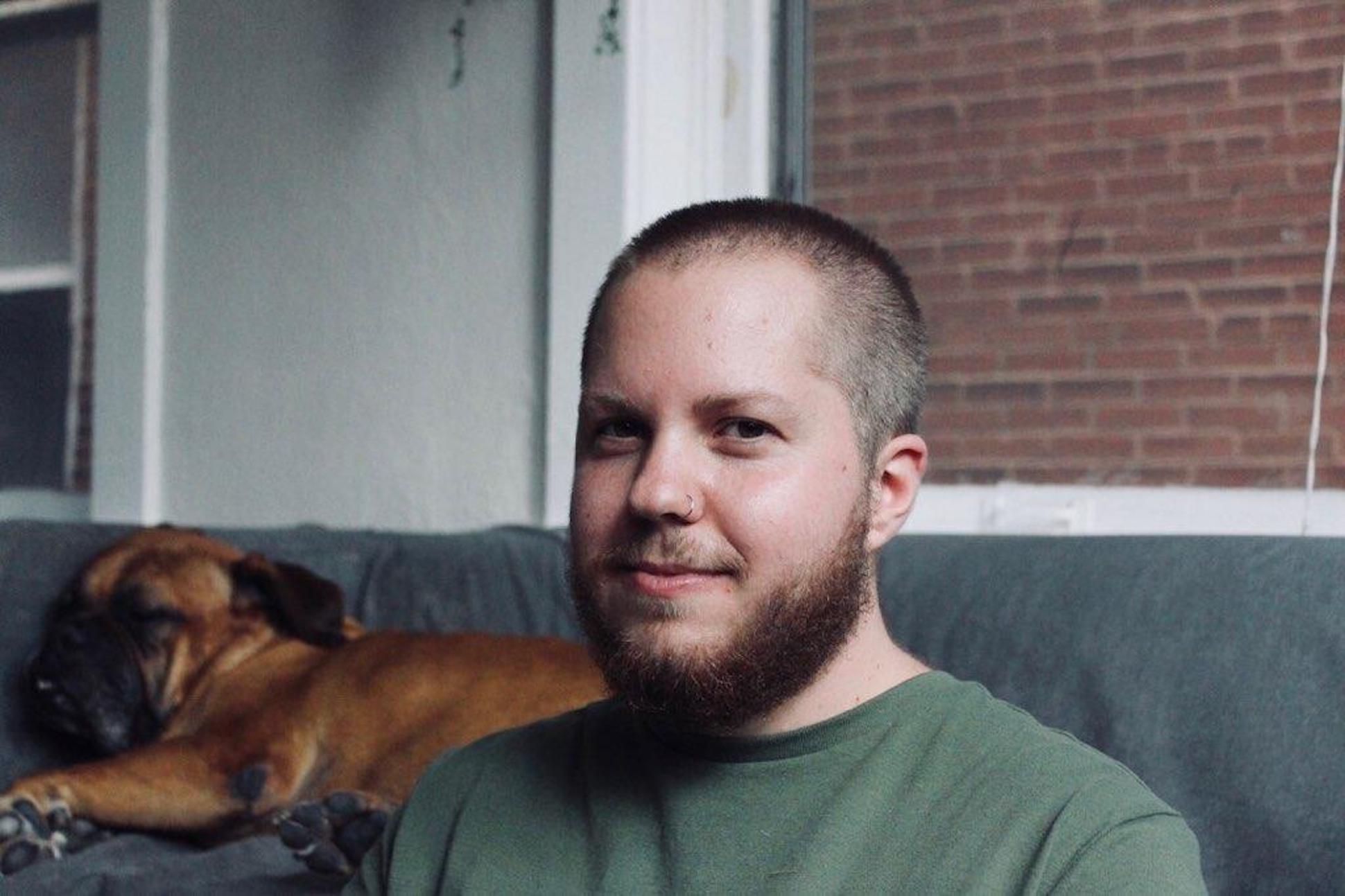 A skinhead white male with beard in a green t-shirt sitting beside a couch where a dog is lying down