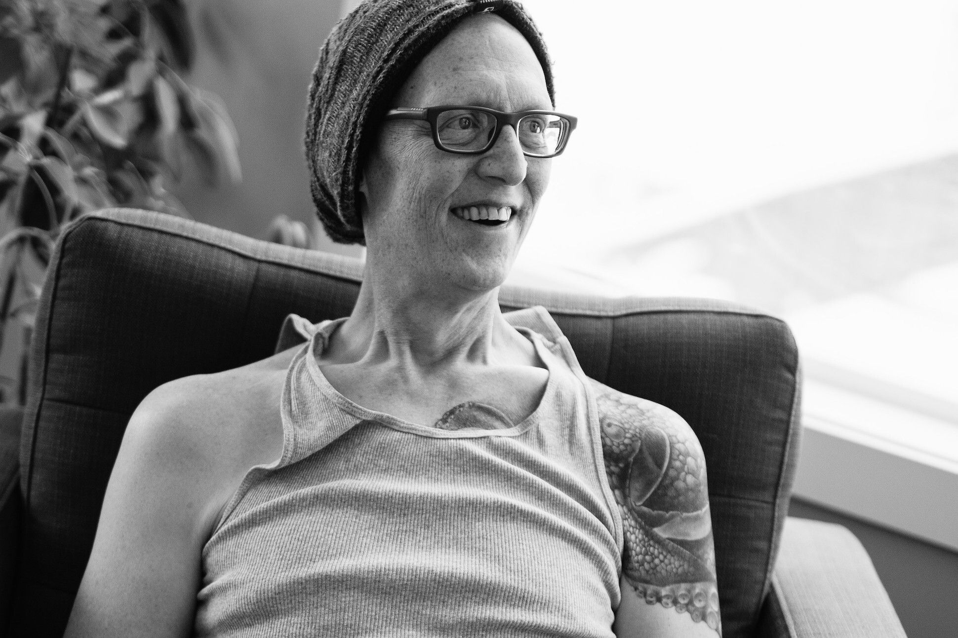 A spectacled man in a beanie and a tank top with tattoo in one arm sitting in an armchair